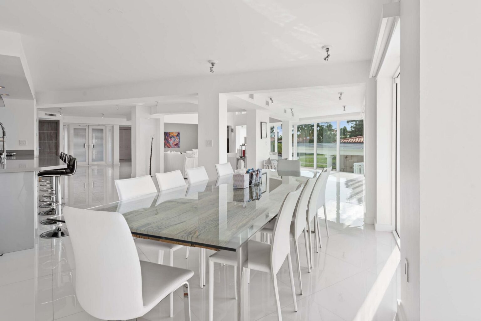 Bright dining space featuring a large glass dining table with seating for eight, tile flooring, expansive windows, contemporary lighting, and an open-concept layout connecting the kitchen, living room, and outdoor entertaining areas.