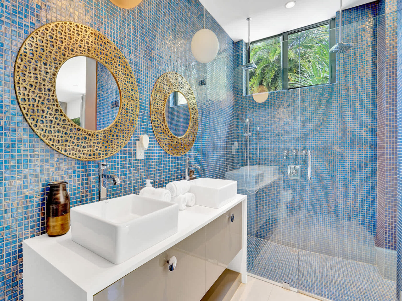 Modern ensuite bathroom with a double vanity, dual vessel sinks, and walk-in glass shower. Decorative blue tile walls, large statement mirrors, and natural light from expansive windows create a bright and functional space.