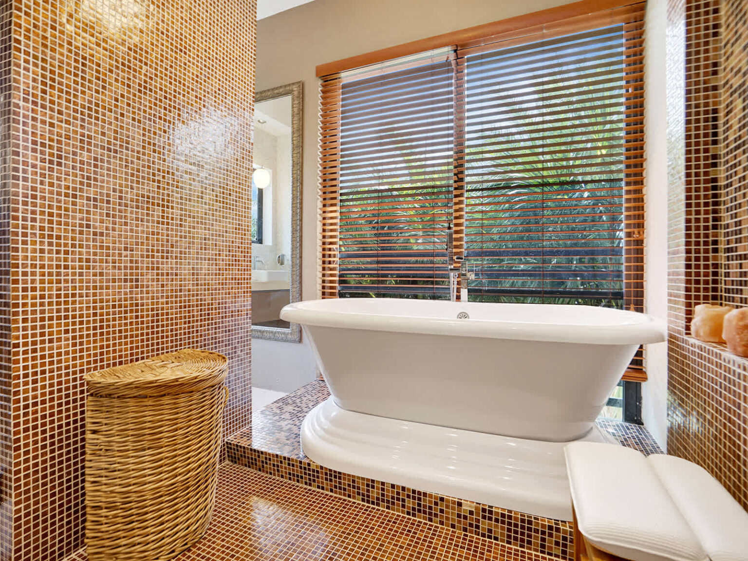 Freestanding soaking tub set within a spa-inspired bathroom featuring mosaic tile accents, natural light, and large windows with plantation shutters. Contemporary finishes and an open layout create a bright and relaxing retreat.