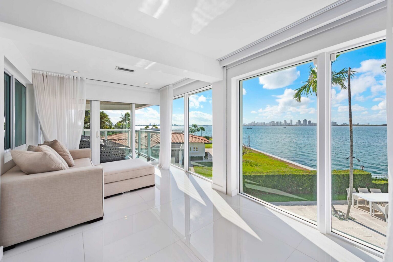 Waterfront sitting area featuring floor-to-ceiling windows, a sectional sofa, tile flooring, and panoramic bay views. Bright open space connects to a private balcony and showcases the water, shoreline, and city skyline beyond.