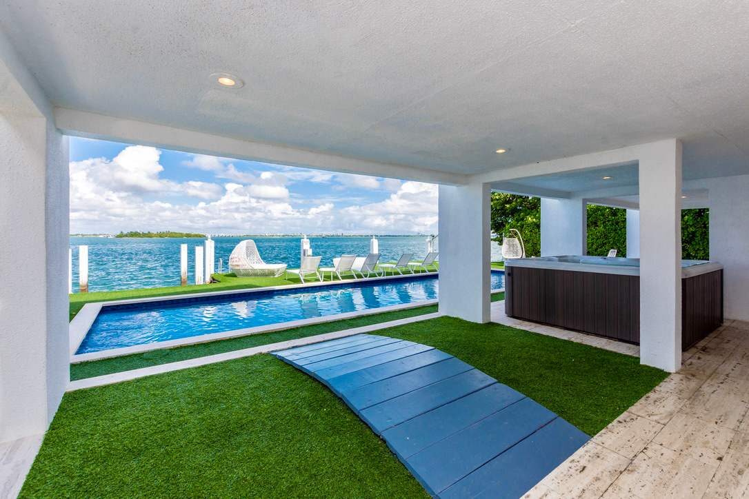 Covered waterfront patio featuring a private swimming pool, artificial turf lounge area, outdoor seating space, open-air entertaining zone, built-in bar counter, glass safety fencing, and panoramic bay views with direct access to poolside amenities.