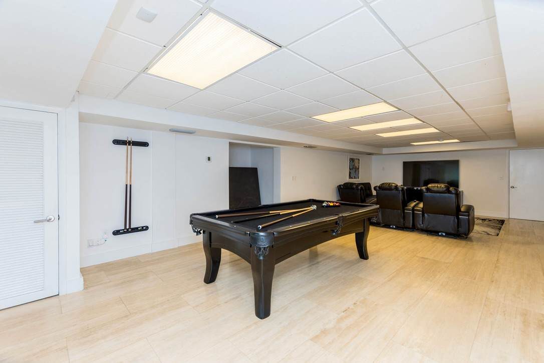 Game and media room featuring a full-size pool table, theater-style recliner seating, large flat-screen TVs, wood-look flooring, suspended ceiling lighting, and a spacious open layout designed for indoor entertainment and group gatherings.