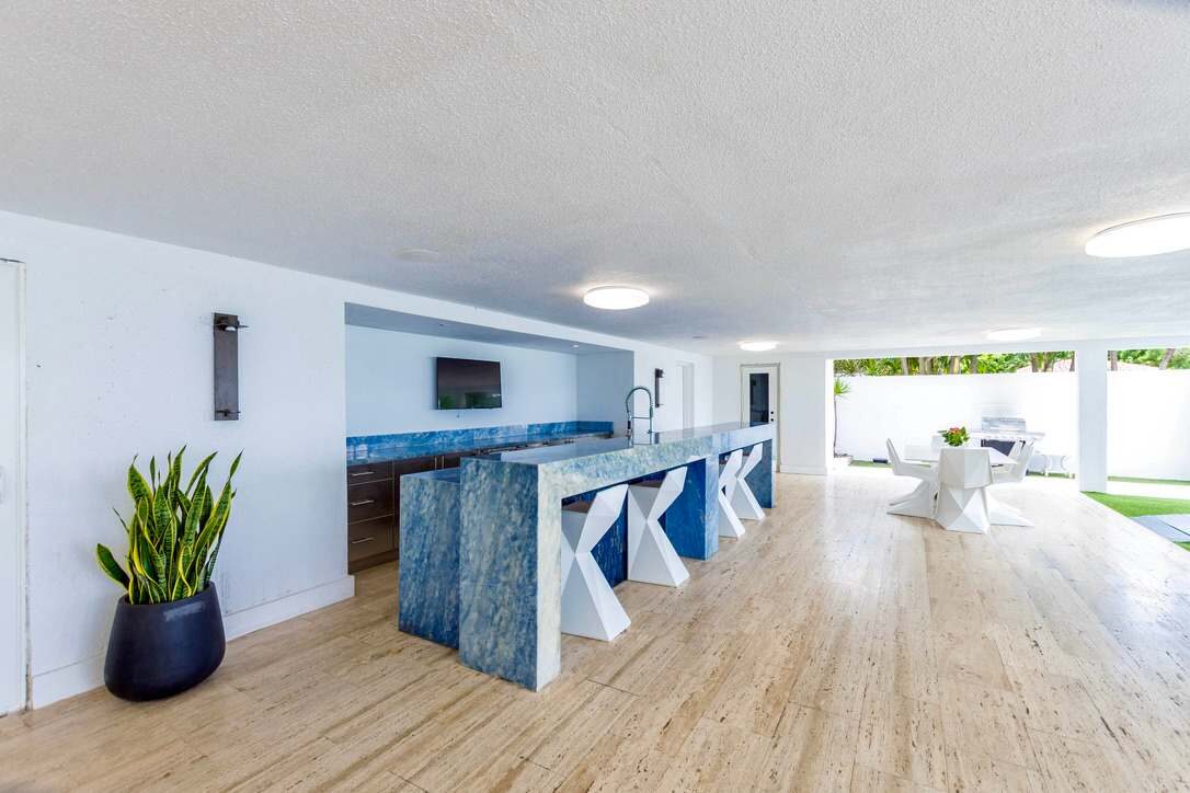 Covered outdoor entertainment area featuring a built-in bar with stone countertop, bar seating, wall-mounted TV, dining space, wood-look tile flooring, open-air layout, and direct access to the pool terrace and landscaped grounds.