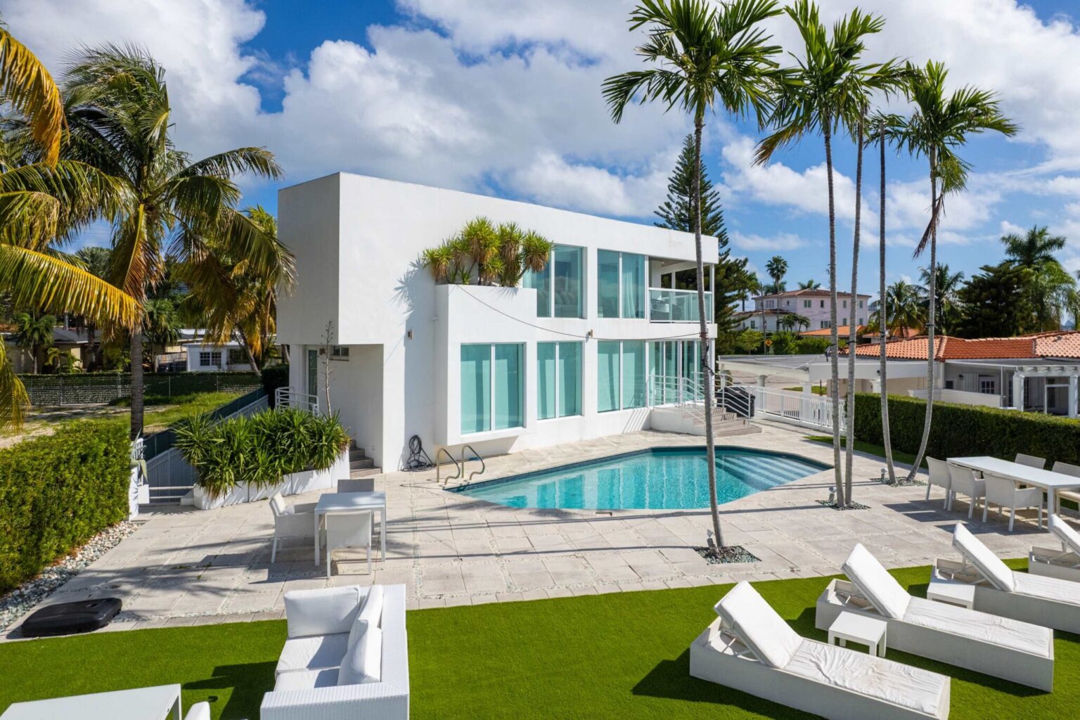 Contemporary waterfront estate featuring a private swimming pool, expansive sun deck, outdoor lounge seating, manicured landscaping, mature palm trees, modern architecture, and direct access to scenic waterfront surroundings.