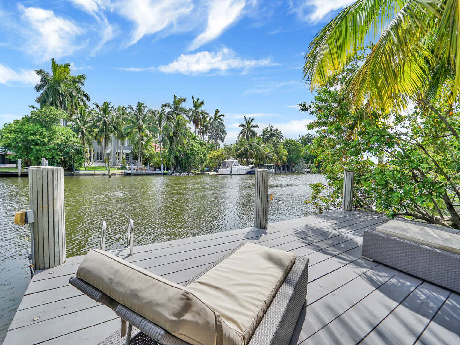 Waterfront deck with cushioned lounge chairs and expansive canal views, surrounded by tropical palm trees and lush greenery. This outdoor relaxation area offers direct water access and a peaceful setting for enjoying the scenery.