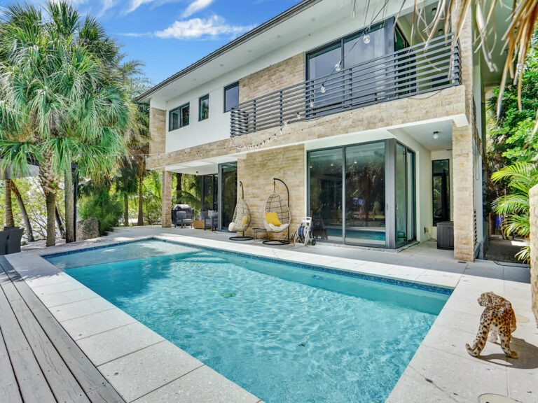 Contemporary two-story villa overlooking a rectangular swimming pool with a tiled deck and poolside seating. Floor-to-ceiling glass doors connect indoor and outdoor living spaces, framed by tropical landscaping and palm trees.