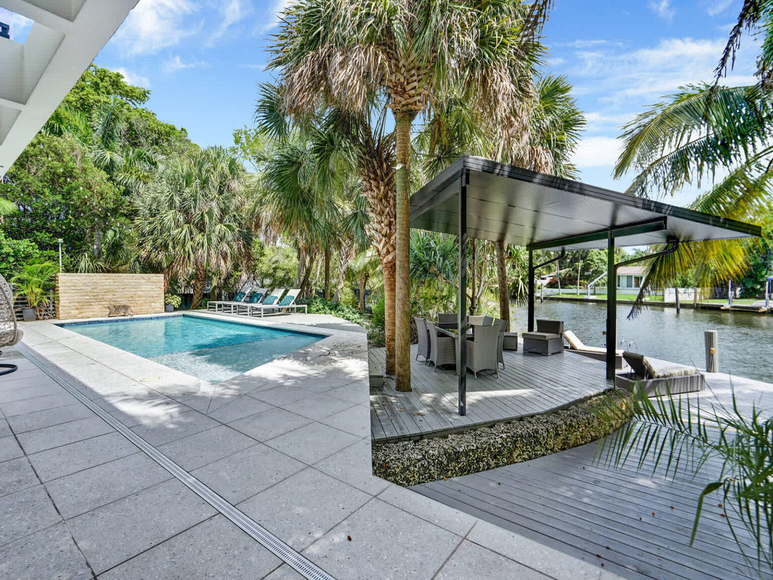 Poolside terrace featuring a modern covered pavilion with outdoor seating, surrounded by tropical landscaping and mature palm trees. Located beside the swimming pool, this spacious area provides shaded space for relaxing and socializing.