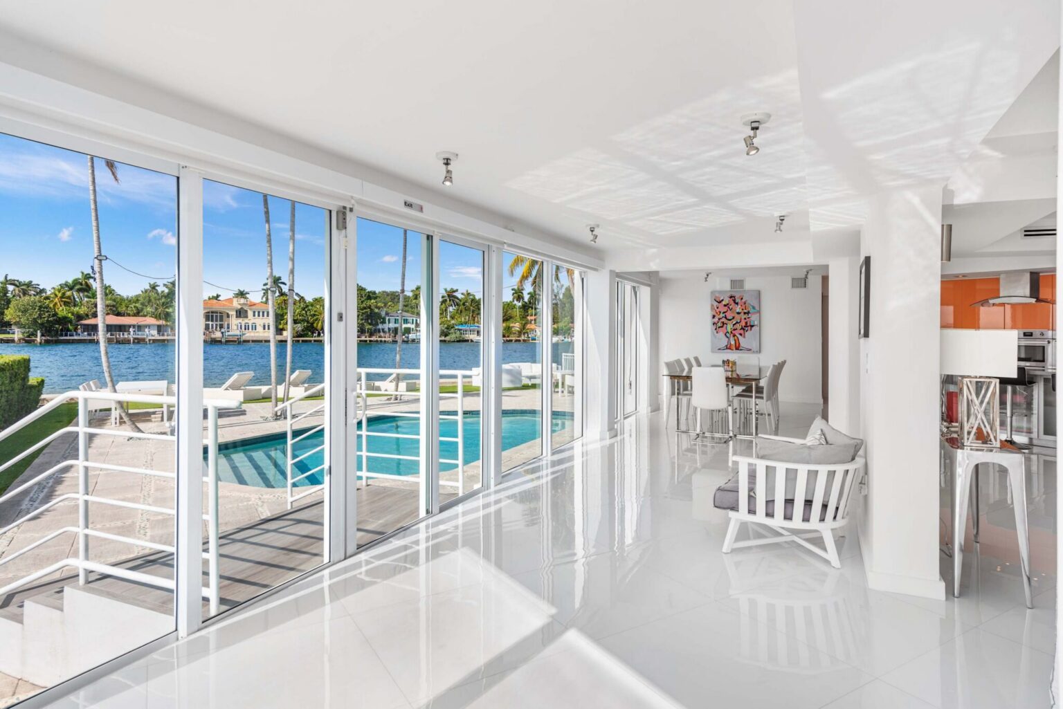 Glass-enclosed waterfront lounge with floor-to-ceiling sliding doors, panoramic water views, direct pool access, glossy tile flooring, bar seating, abundant natural light, and seamless indoor-outdoor living space.