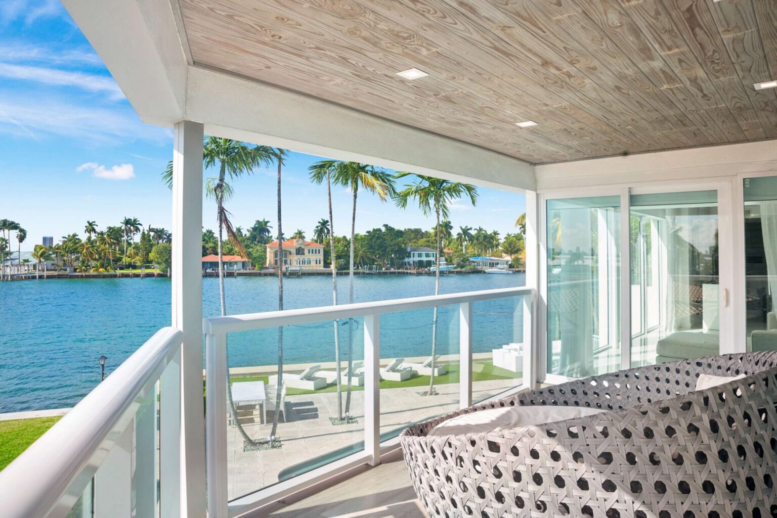 Bayfront balcony lounge featuring cushioned outdoor seating, glass railings, wood-look ceiling finishes, and panoramic water views. Covered outdoor space overlooks the bay, private lawn, pool area, and neighboring waterfront residences.