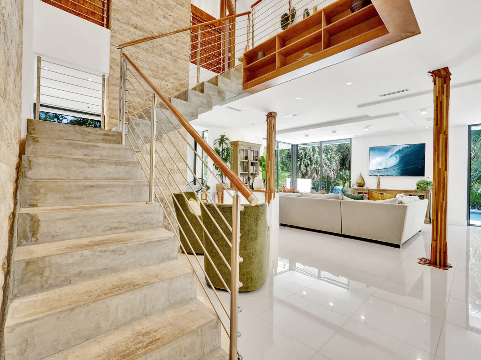 Open-concept interior showcasing a floating staircase with wood and metal railings, polished tile flooring, and natural stone accents. Large windows provide abundant natural light, connecting the living, dining, and lounge areas.
