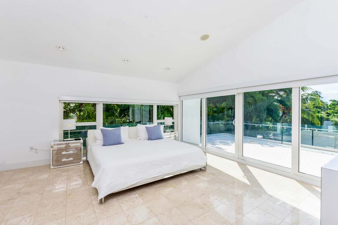 Sun-filled primary bedroom featuring a king-size bed with premium linens, floor-to-ceiling sliding glass doors, private balcony access, tile flooring, mirrored nightstands, recessed lighting, and expansive windows overlooking lush tropical greenery.