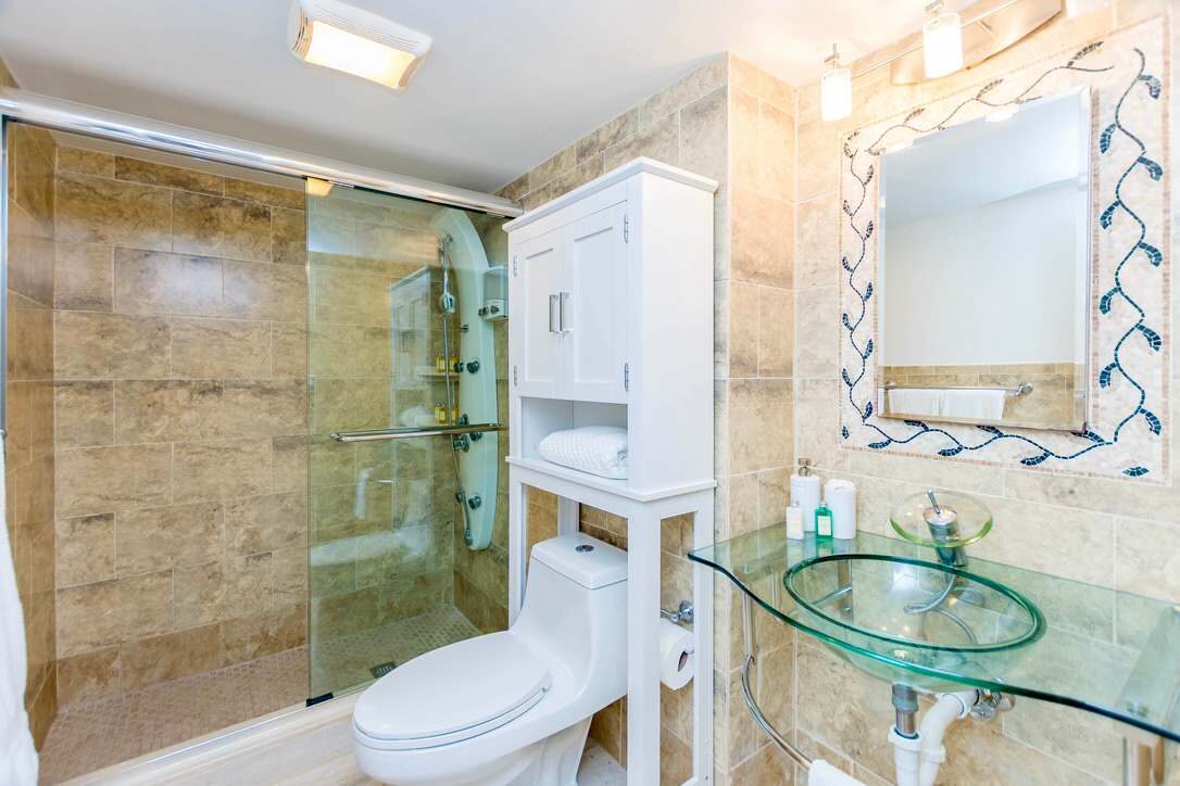 Full guest bathroom featuring a glass-enclosed walk-in shower, natural stone tile finishes, glass vessel sink, decorative framed mirror, storage cabinet, modern fixtures, and a functional layout with dedicated counter space and built-in shelving.