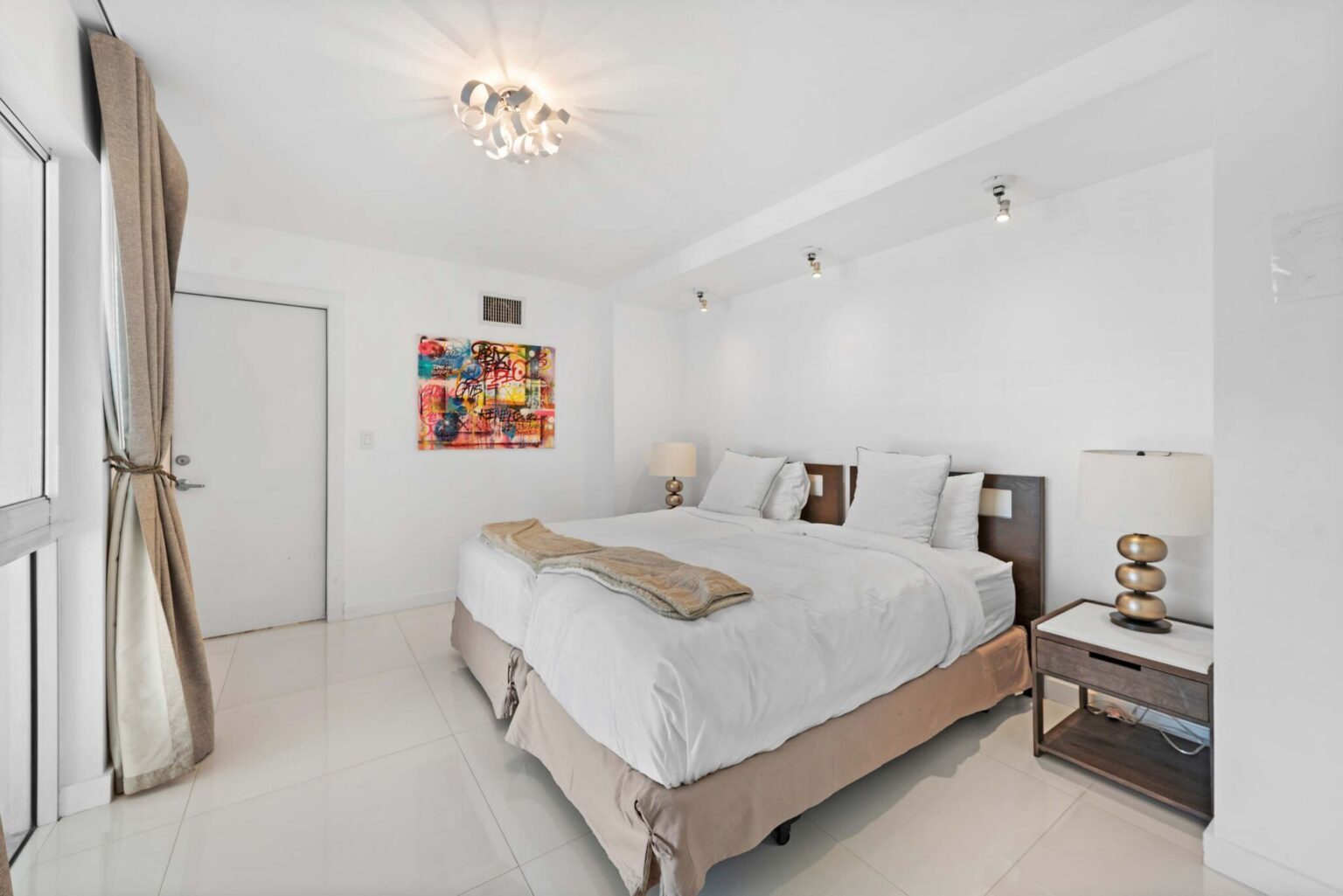 Modern guest bedroom featuring a queen-size bed with white linens, bedside tables with lamps, large windows with blackout curtains, contemporary artwork, overhead lighting, and a bright, minimalist layout designed for comfort and extended stays.