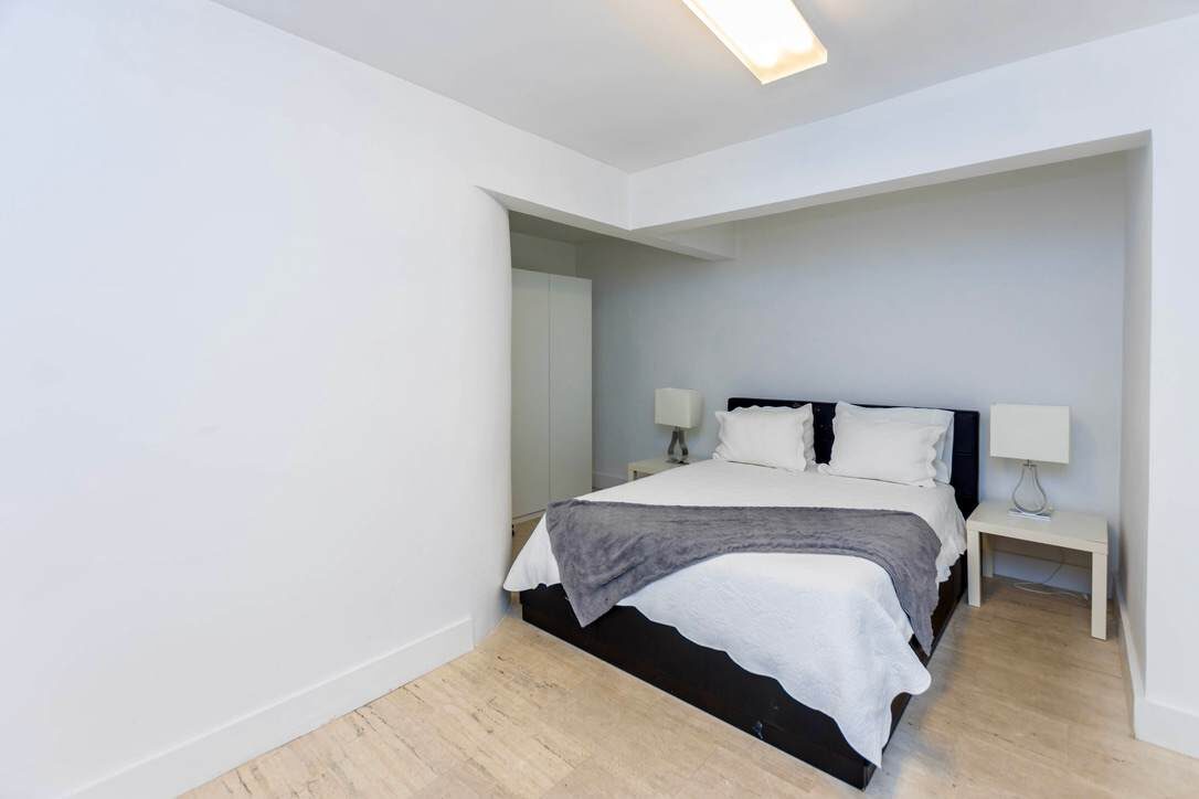 Cozy guest bedroom featuring a queen-size bed with upholstered frame, bedside tables with lamps, wood-look flooring, recessed sleeping alcove, overhead lighting, and a clean minimalist design with convenient access to adjacent living spaces.