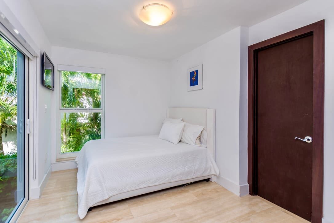 Private guest bedroom featuring a queen-size bed with white linens, large windows with garden views, tile flooring, wall-mounted TV, bedside lighting, and a bright minimalist layout with direct access to outdoor spaces.