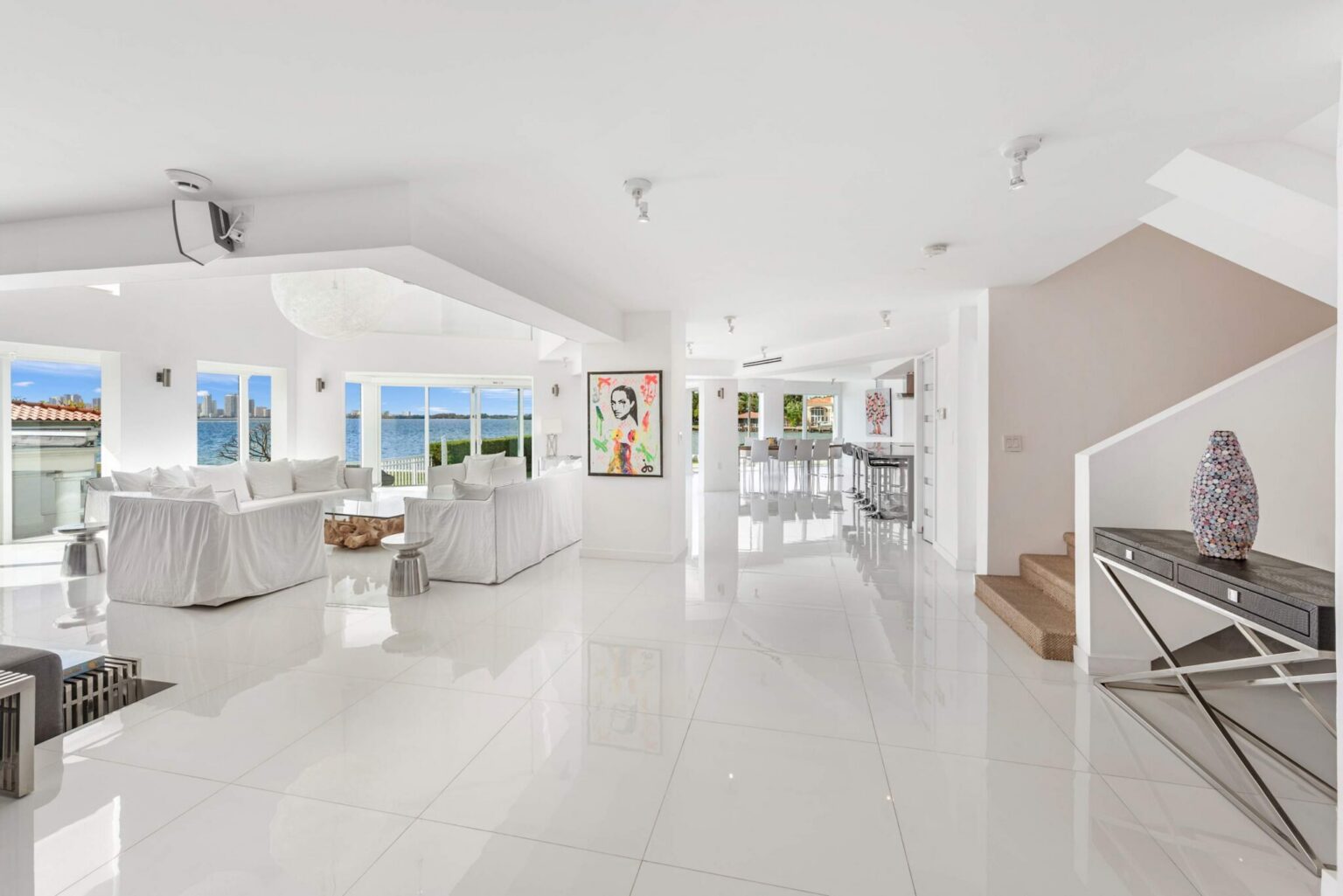 Expansive open-concept main level featuring glossy tile flooring, waterfront living areas, contemporary artwork, staircase access, abundant natural light, and seamless connectivity between the media lounge, kitchen, dining, and gathering spaces.