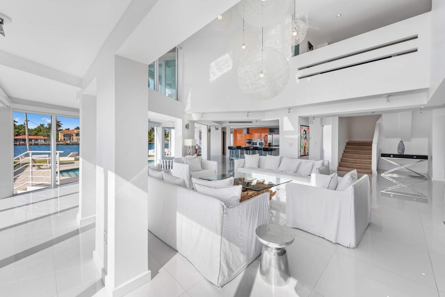 Double-height living area with floor-to-ceiling windows, white sectional seating, modern chandelier, open-concept design, waterfront views, glossy tile flooring, and direct sightlines to the kitchen and upper-level loft space.