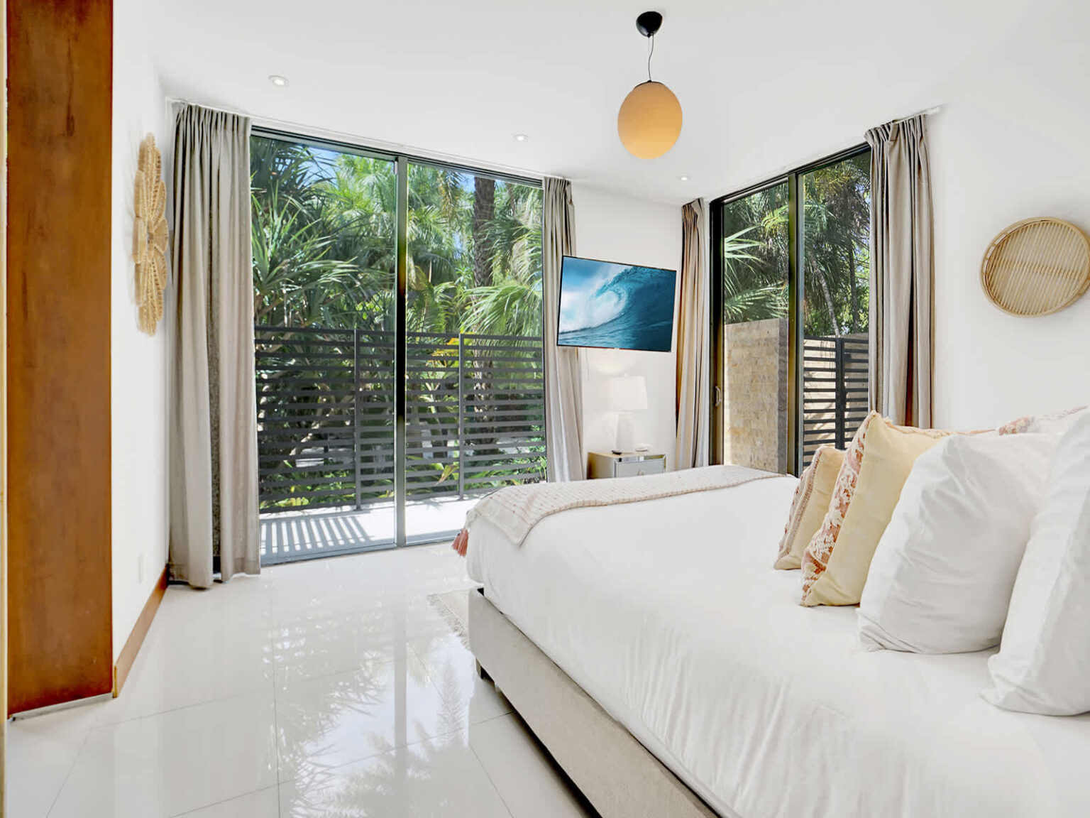 Bright bedroom featuring a king-size bed, floor-to-ceiling corner windows, and sliding glass doors with tropical garden views. Natural light fills the space, while modern furnishings, blackout curtains, and clean finishes create a comfortable retreat