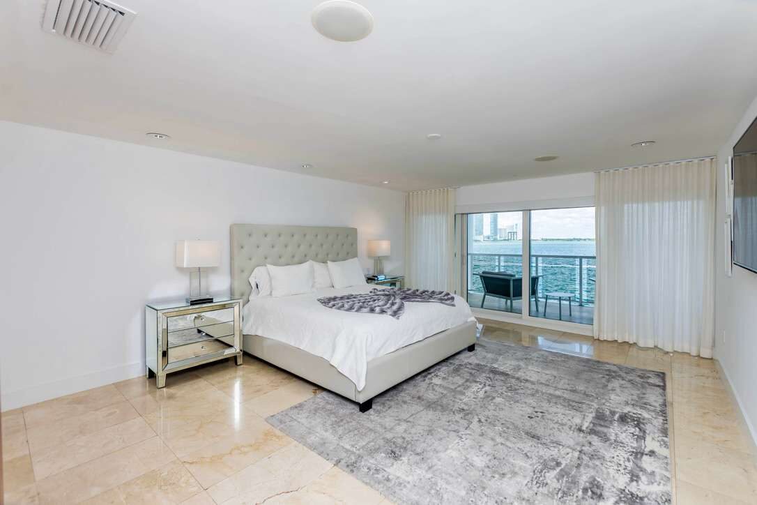 Spacious primary suite featuring a king-size bed with upholstered headboard, premium linens, mirrored nightstands, area rug, tile flooring, recessed lighting, and sliding glass doors leading to a private balcony with water views.