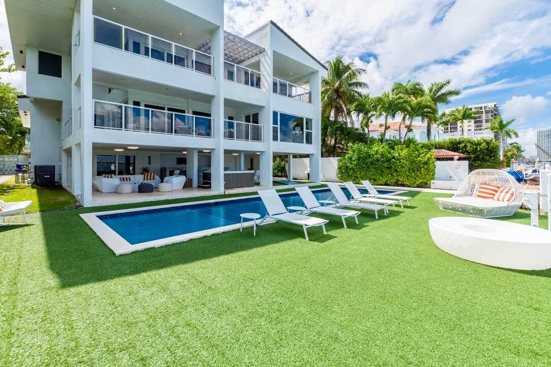 Bayfront villa exterior featuring a private swimming pool, poolside sun loungers, expansive artificial turf lawn, covered outdoor living area, multiple balconies, floor-to-ceiling windows, tropical landscaping, and direct water access.