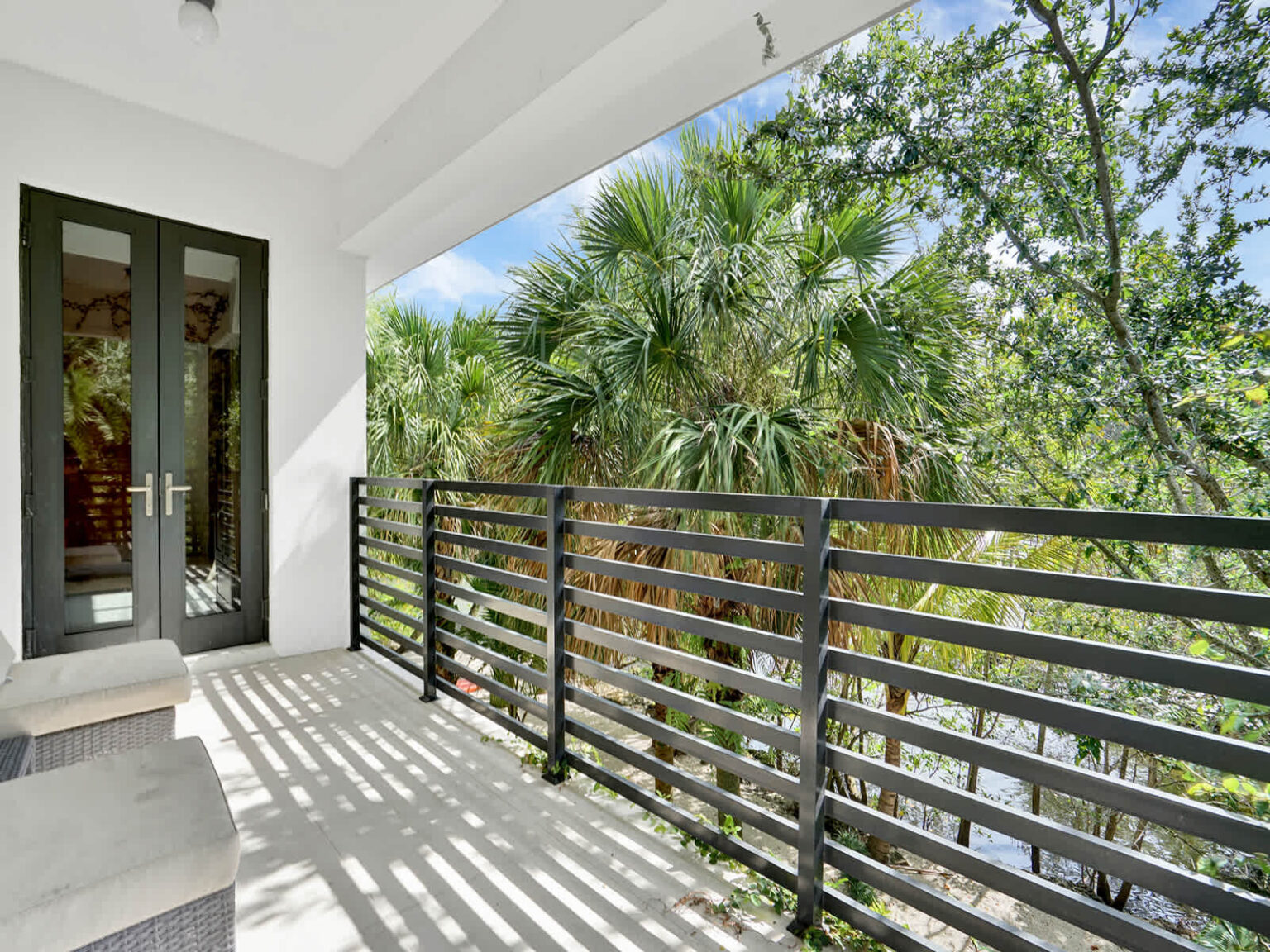 Covered second-floor balcony with modern metal railings and elevated views of tropical palm trees and lush greenery. This outdoor space provides fresh air, natural light, and a peaceful setting that extends the adjacent bedroom.