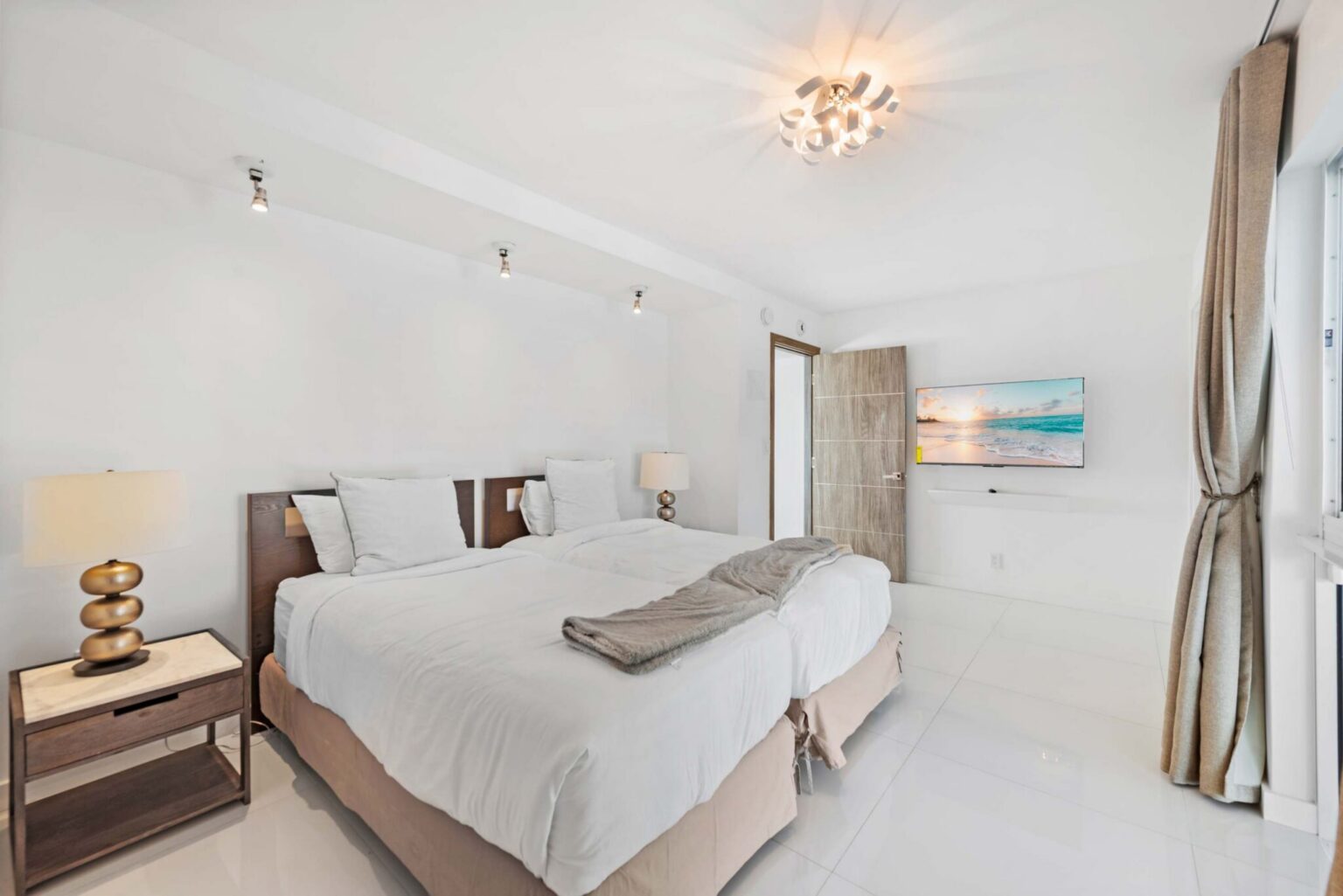 Inviting guest bedroom featuring a queen-size bed with white linens, bedside tables with lamps, large window with blackout curtains, built-in closet storage, modern lighting, and a bright, functional layout designed for comfort and extended stays.