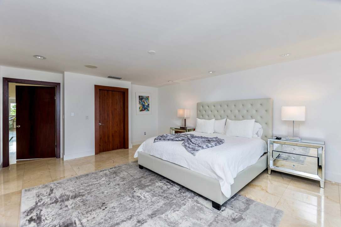 Oversized primary bedroom featuring a king-size bed with upholstered headboard, mirrored nightstands, premium linens, area rug, tile flooring, recessed lighting, and a spacious open layout with direct access to the suite's private amenities.