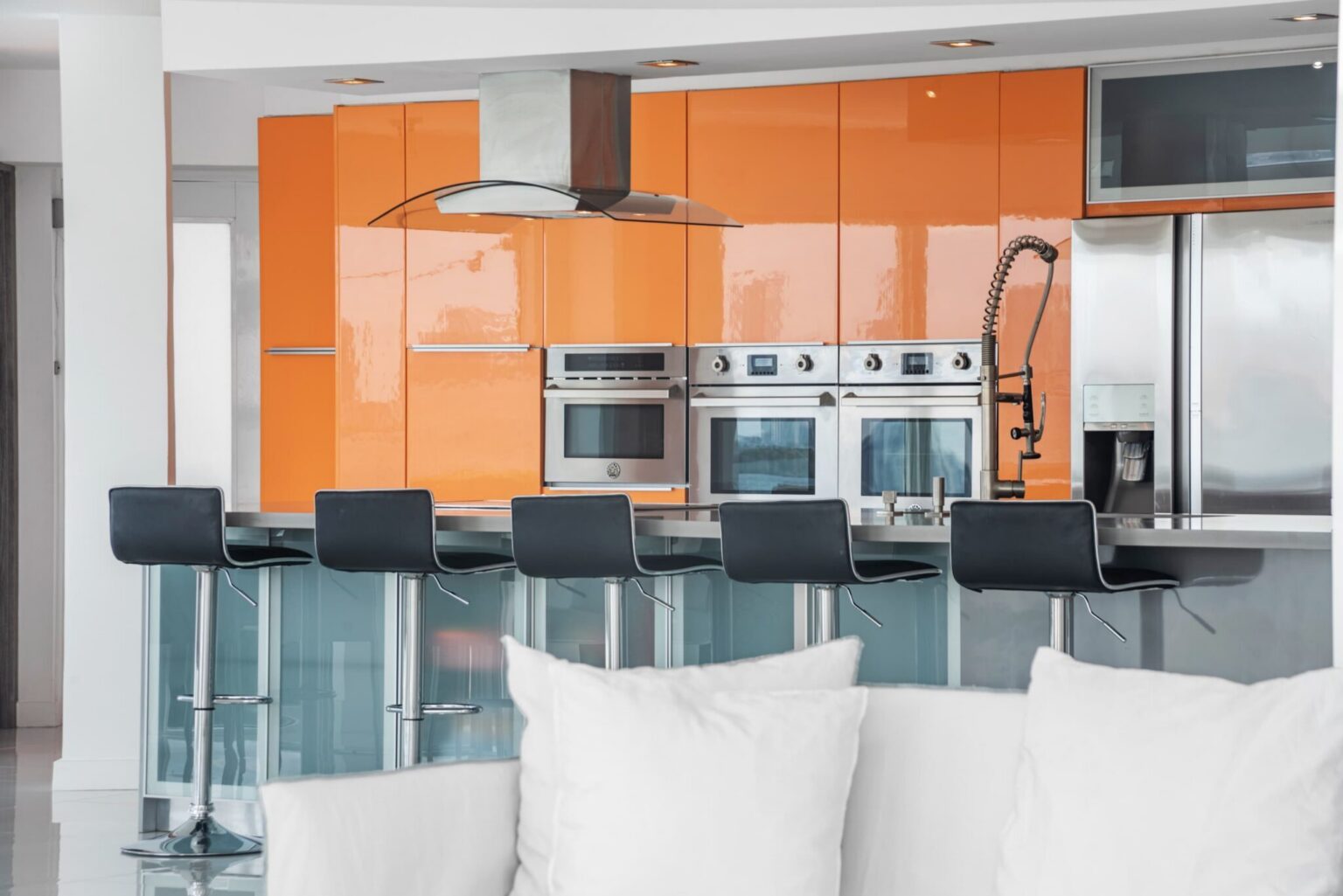 Modern kitchen featuring glossy orange cabinetry, stainless steel appliances, double wall ovens, French-door refrigerator, glass breakfast bar with seating for six, built-in microwave, and open-concept layout overlooking the living area.