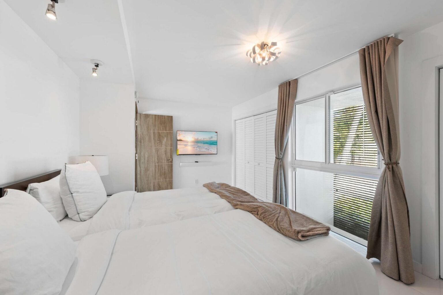 Twin guest bedroom featuring two twin-size beds with white linens, a large window with blackout curtains, built-in closet storage, wood-look flooring, modern lighting, and a bright layout designed for flexible sleeping arrangements.