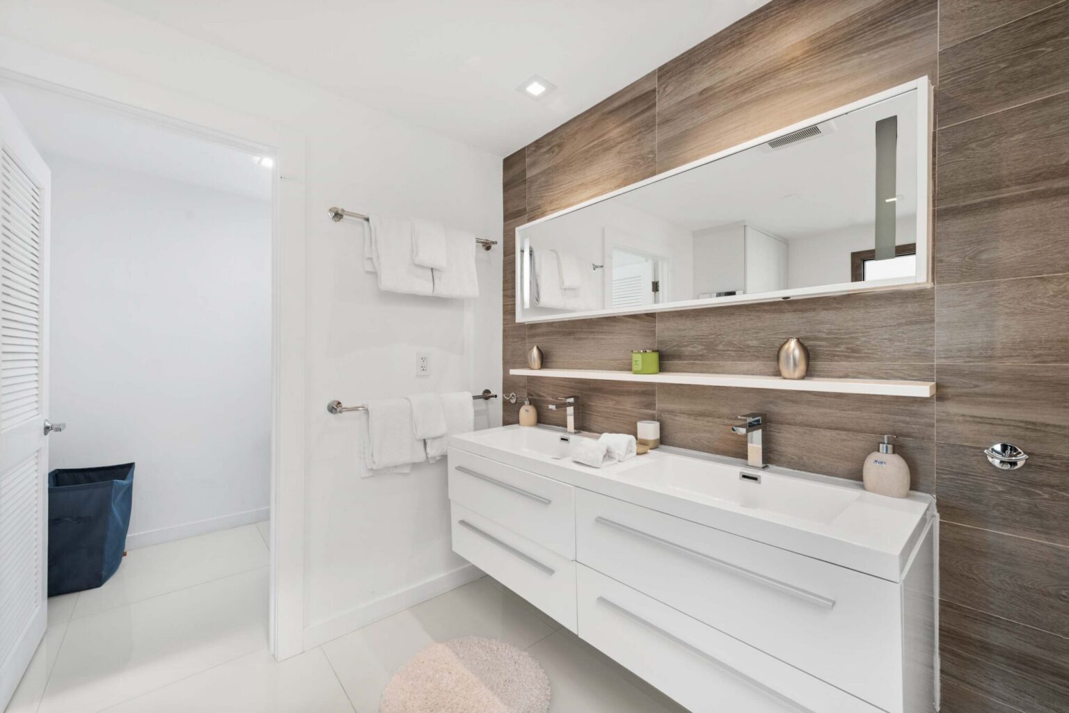 Double-vanity bathroom featuring dual sinks, a floating white vanity with storage drawers, oversized wall mirror, wood-look accent tile, modern fixtures, towel storage, and a bright contemporary design with ample counter space for guests.