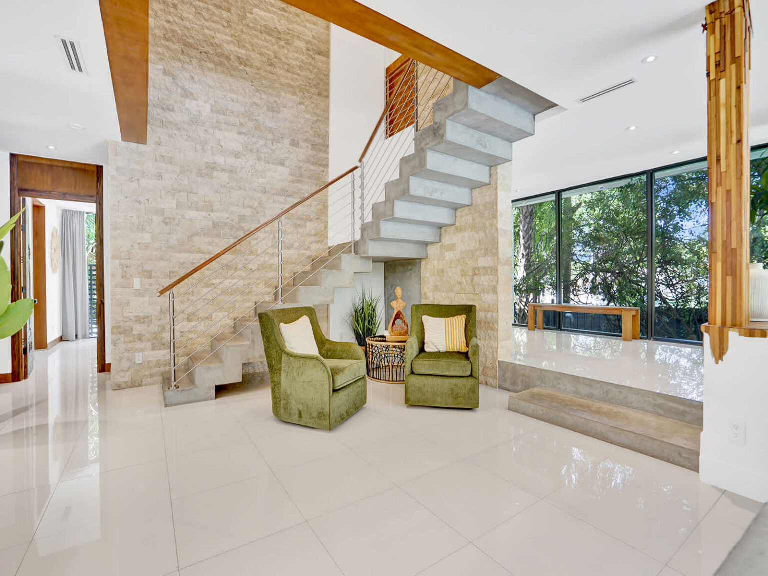 Architectural staircase with floating concrete steps, natural wood accents, and a stone feature wall. This open interior space includes comfortable lounge seating, polished tile floors, and large windows overlooking tropical greenery.