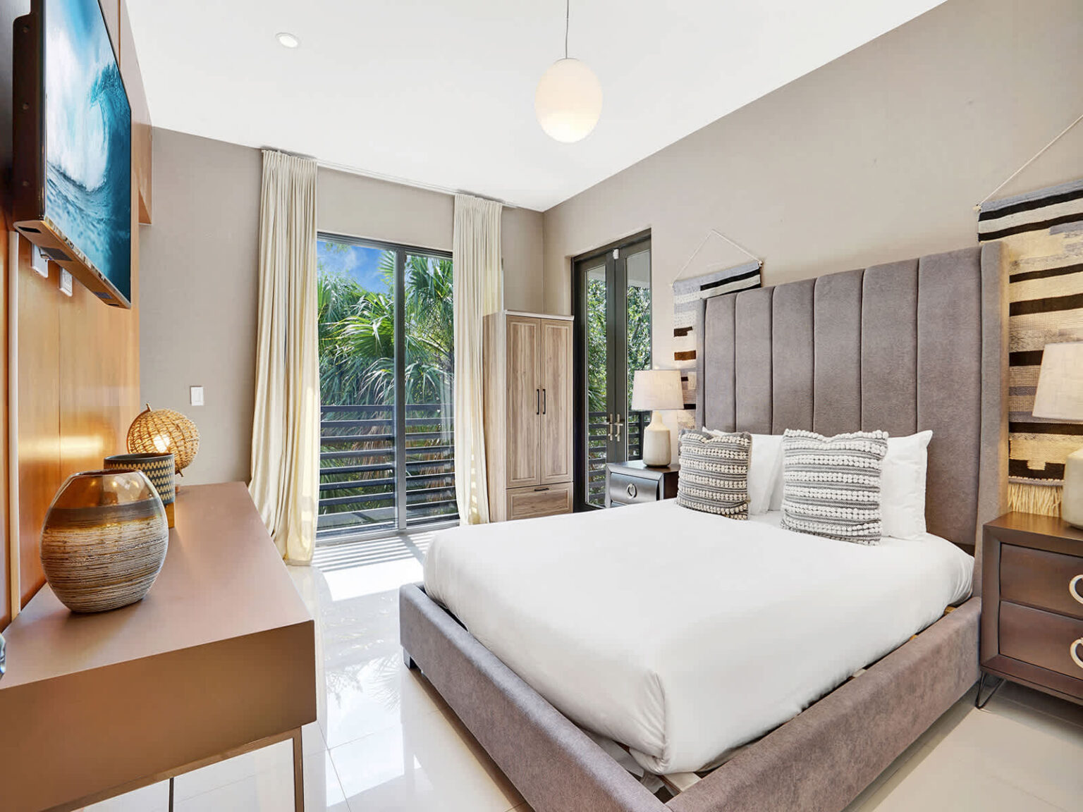 Comfortable bedroom featuring a king-size bed, upholstered headboard, and sliding glass doors with tropical views. Natural light, modern furnishings, and a bright open layout create a relaxing retreat with indoor-outdoor connectivity.