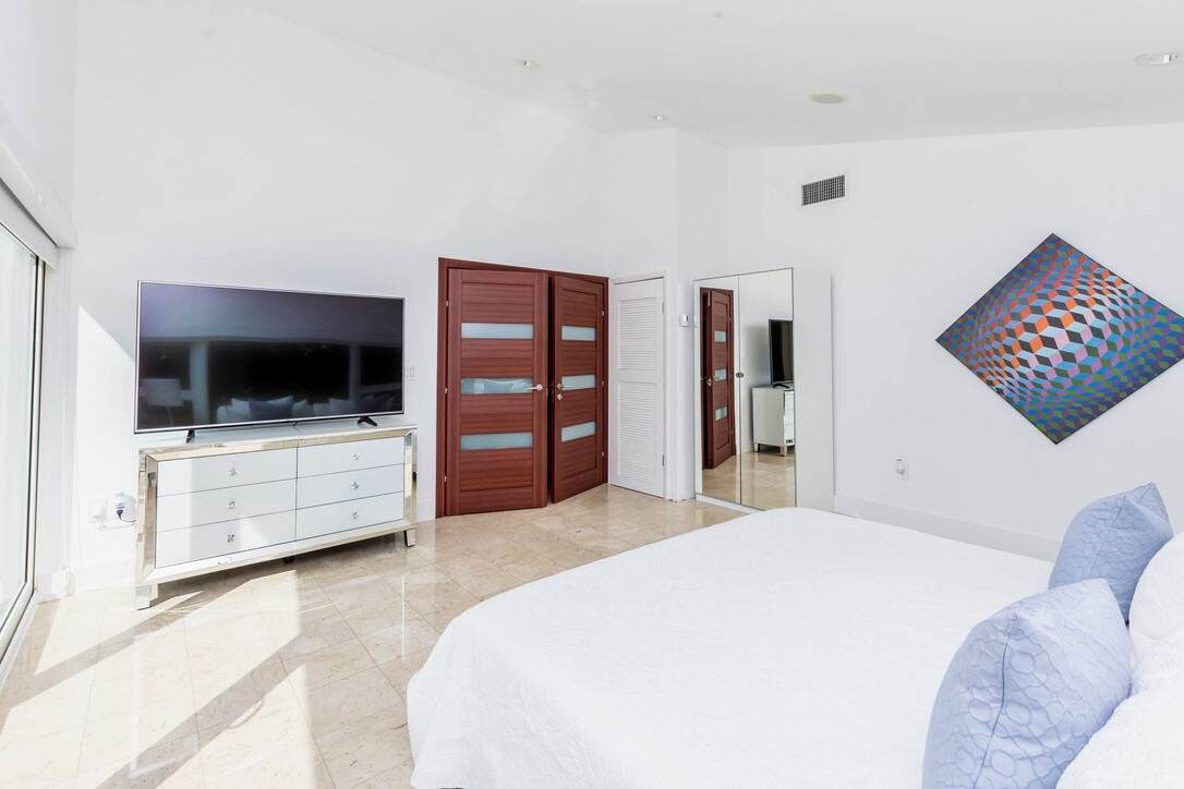 Primary bedroom suite featuring a king-size bed with premium linens, wall-mounted smart TV, dresser storage, tile flooring, contemporary artwork, recessed lighting, and sliding glass doors that bring in abundant natural light.