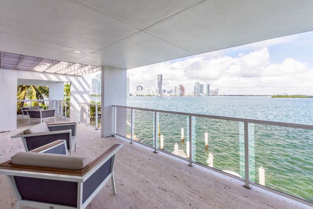 Bayfront balcony retreat featuring cushioned outdoor lounge seating, glass railings, wood-look flooring, and expansive water views. Covered outdoor space provides direct access from the villa and overlooks the bay, skyline, and private dock area.