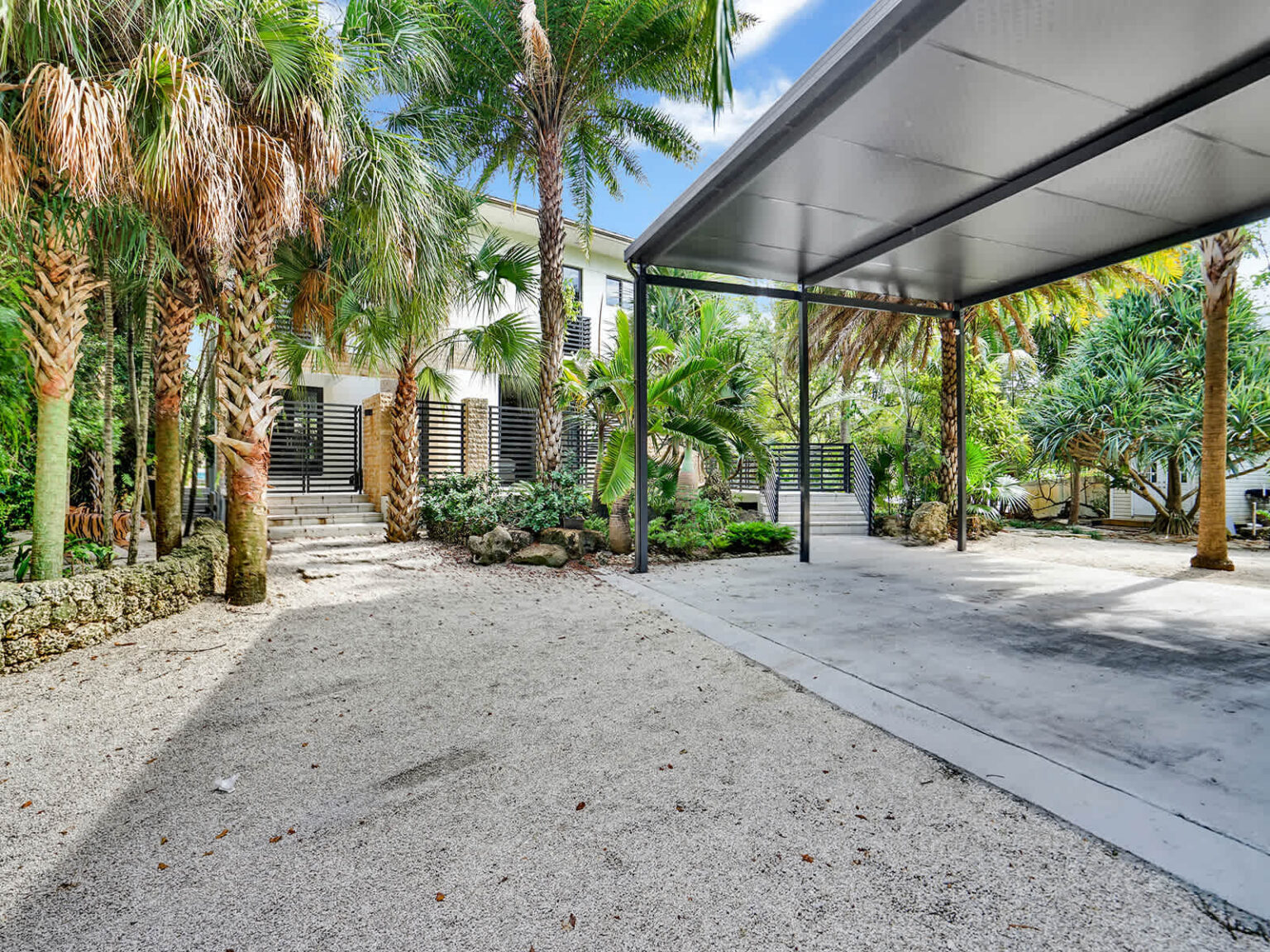 Covered parking area with a metal canopy, spacious concrete driveway, and surrounding tropical landscaping. Mature palm trees and lush greenery create a welcoming arrival experience, with convenient access to the home and outdoor spaces.