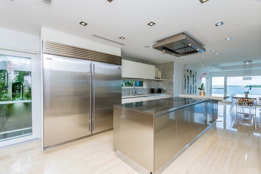 Chef’s kitchen featuring a large stainless steel island, commercial-grade refrigerator, built-in cooktop with ceiling-mounted range hood, custom cabinetry, recessed lighting, and an open-concept layout connected to the dining and living areas.