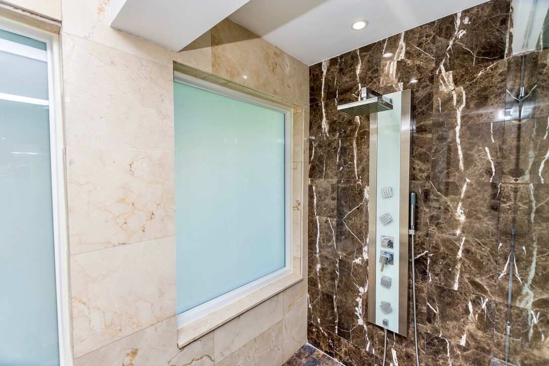 Spa-inspired shower featuring floor-to-ceiling marble-look tile, a glass-enclosed walk-in shower, multi-function body jets, rainfall showerhead, frosted privacy window, recessed lighting, and contemporary fixtures with luxury finishes throughout.