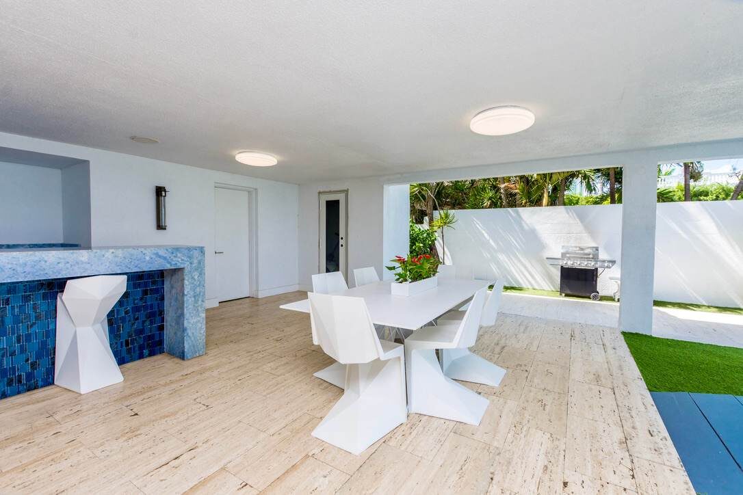 Open-air social area featuring a dining table with seating for six, custom stone bar, covered patio design, wood-look tile flooring, and convenient access to the pool and garden.