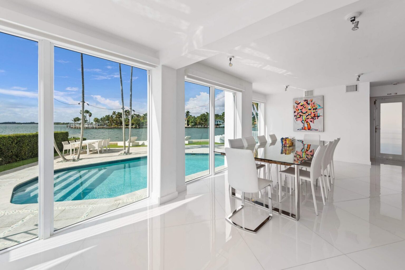 Pool-view dining area featuring a contemporary dining table with seating for six, floor-to-ceiling windows, tile flooring, modern artwork, and an open-concept layout. Bright interior overlooks the private pool and waterfront surroundings.