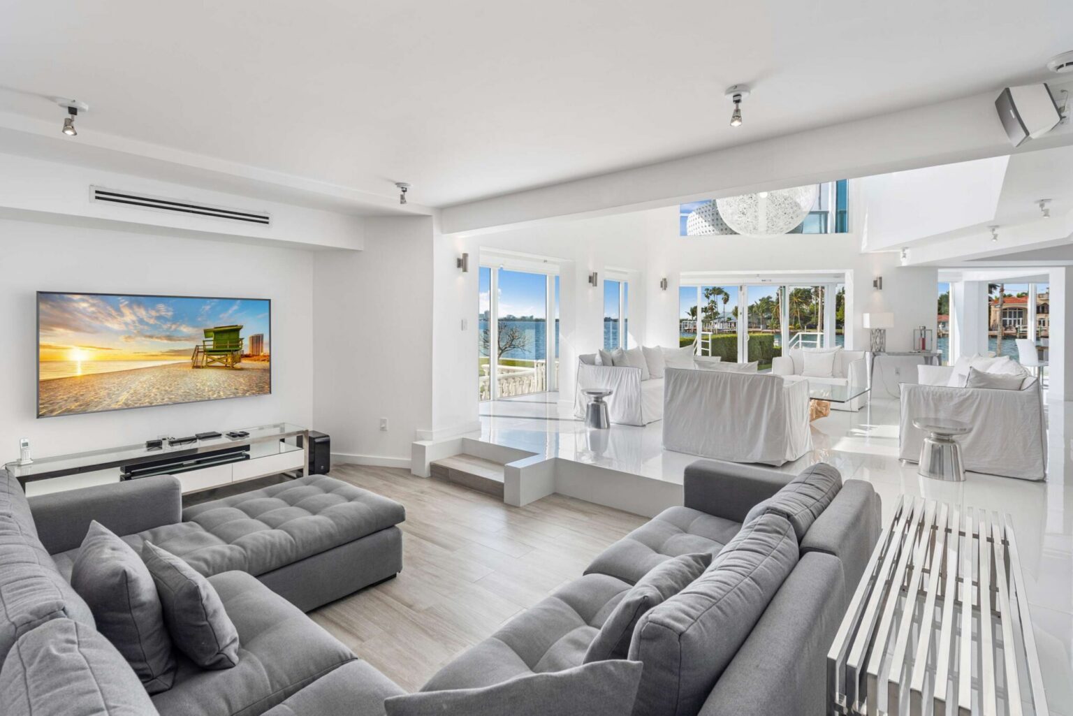 Media lounge with oversized sectional seating, wall-mounted TV, surround sound speakers, light wood flooring, large windows, and an open-concept layout connecting the entertainment area to the main living and waterfront spaces.