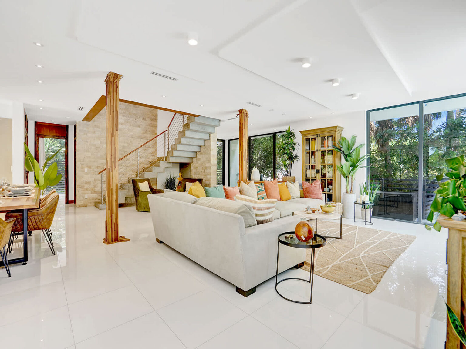 Spacious open-concept living room with modern sectional seating, polished tile flooring, and abundant natural light. Features natural wood columns, a stone accent wall, and seamless access to outdoor living areas and tropical views.