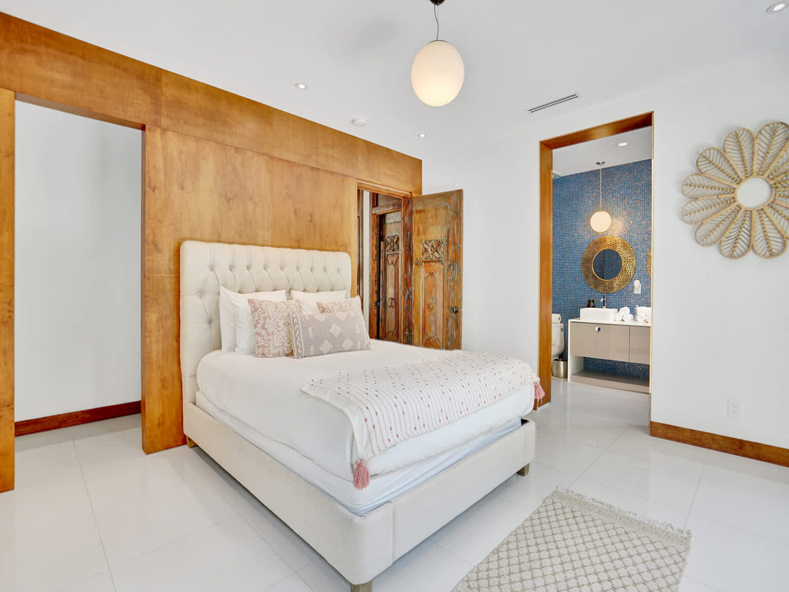 Primary bedroom featuring a king-size upholstered bed, wood accent wall, and modern furnishings. Includes direct access to an ensuite bathroom, polished tile flooring, and a bright, open layout designed for comfort and functionality.