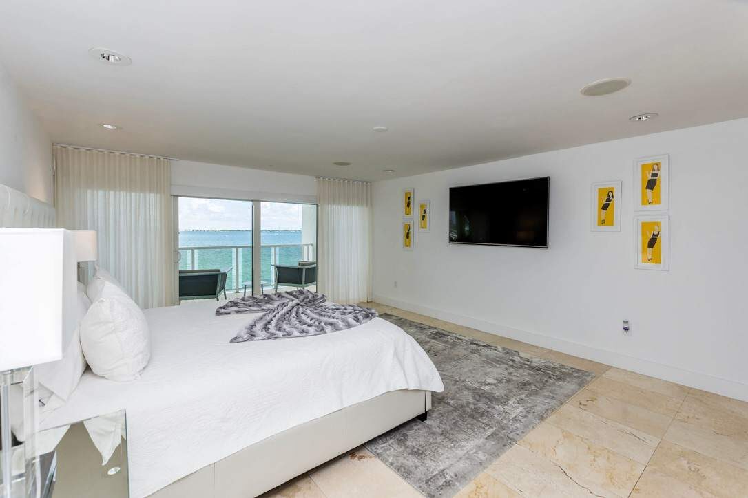 Water-view primary bedroom featuring a king-size bed with premium linens, wall-mounted smart TV, area rug, tile flooring, recessed lighting, and sliding glass doors that open to a private balcony overlooking the bay and surrounding waterfront scenery