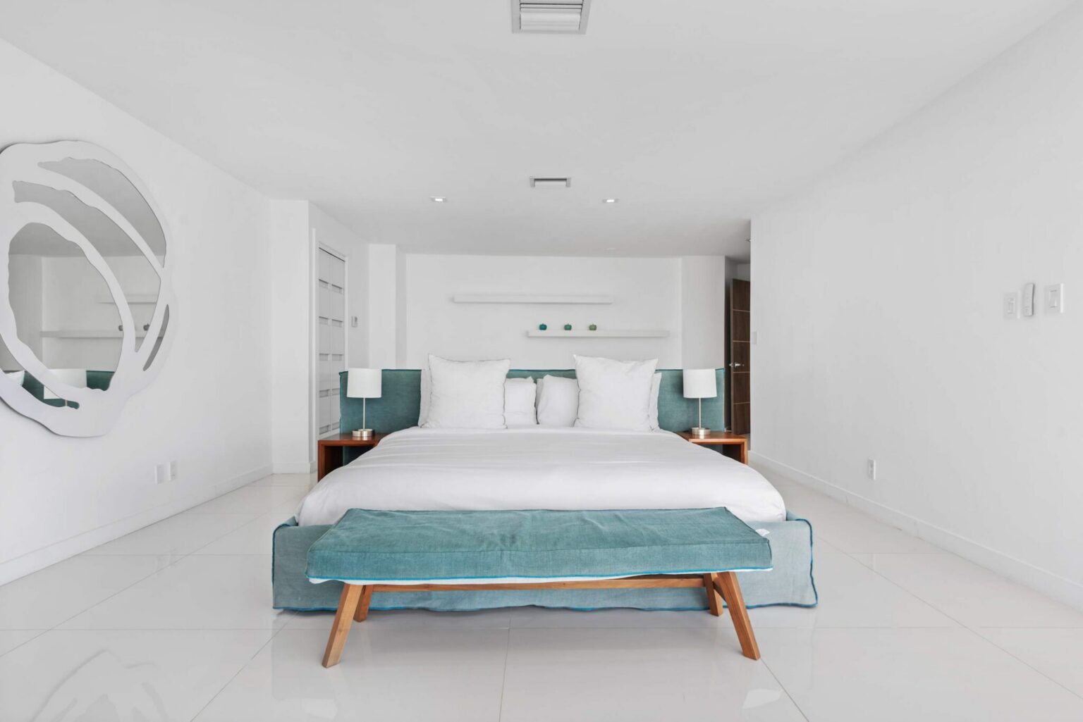 Spacious primary suite featuring a king-size bed with upholstered headboard, bench seating, tile flooring, bedside lamps, built-in shelving, and an open layout designed for comfort, relaxation, and extended stays.
