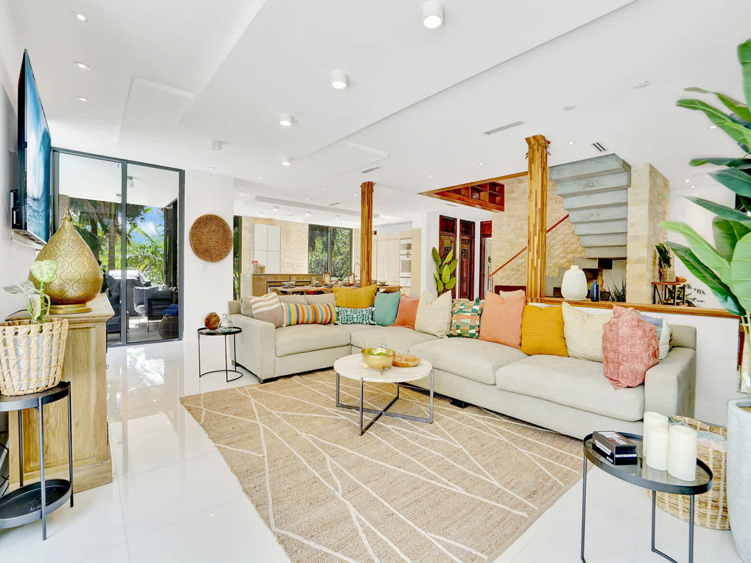 Contemporary living room with a large sectional sofa, area rug, and open-concept layout. Features high ceilings, natural wood columns, abundant natural light, and direct connections to the dining area, staircase, and outdoor spaces.