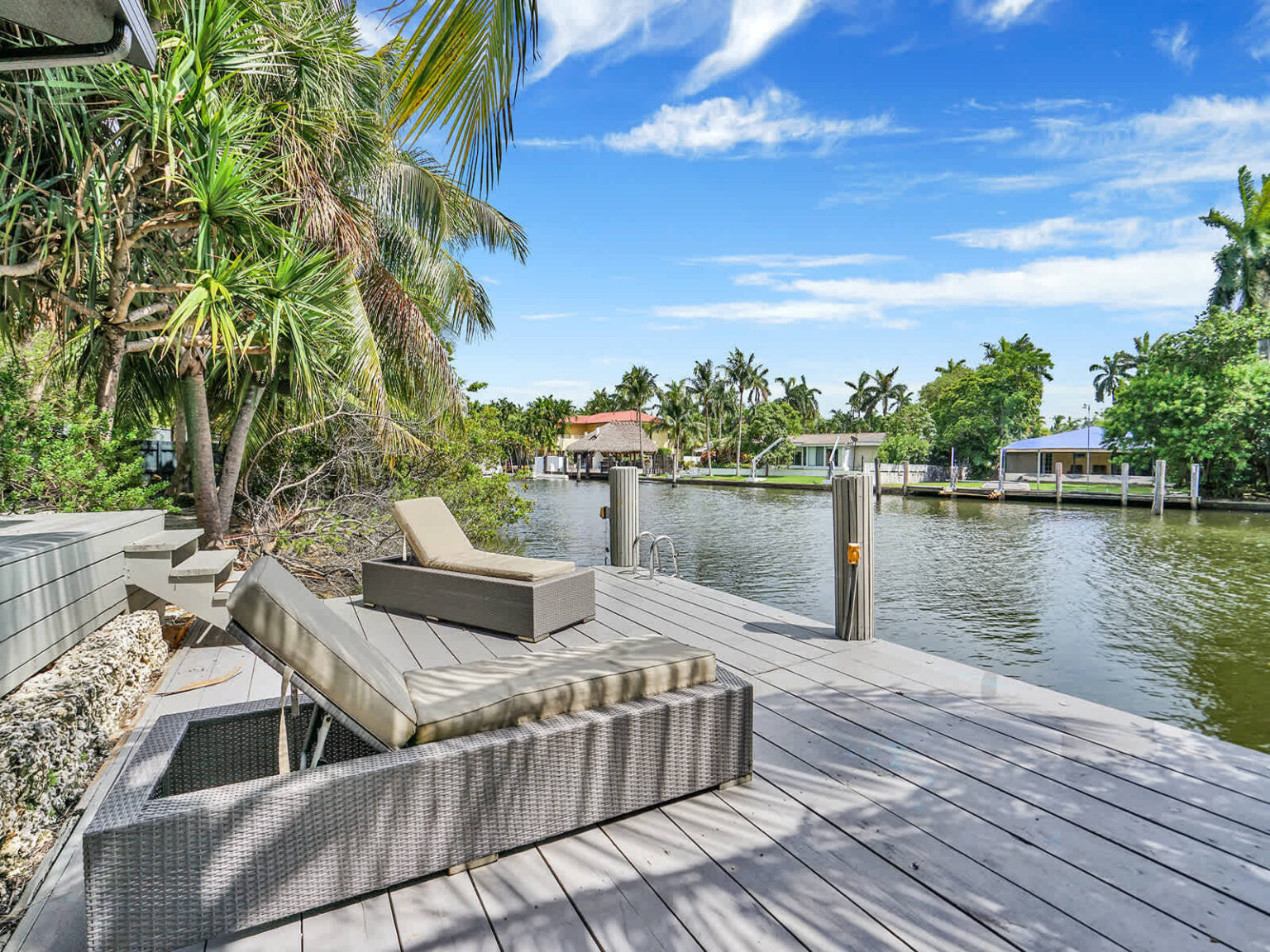 Waterfront deck with cushioned lounge seating and unobstructed canal views. Surrounded by tropical palm trees and landscaped greenery, this outdoor space provides a comfortable setting for relaxing by the water.