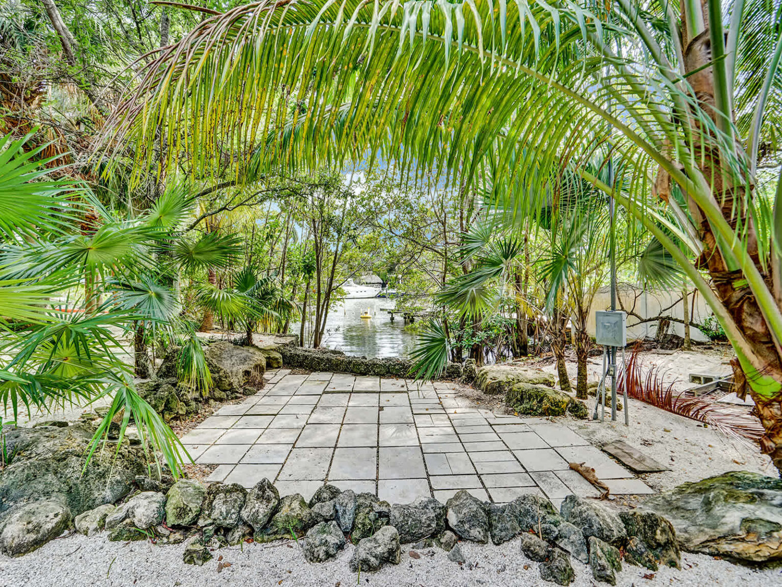 ropical garden pathway framed by arching palm fronds, mature landscaping, and natural stone borders. The paved walkway creates a welcoming entrance experience, surrounded by lush greenery that enhances the property's resort-style setting.