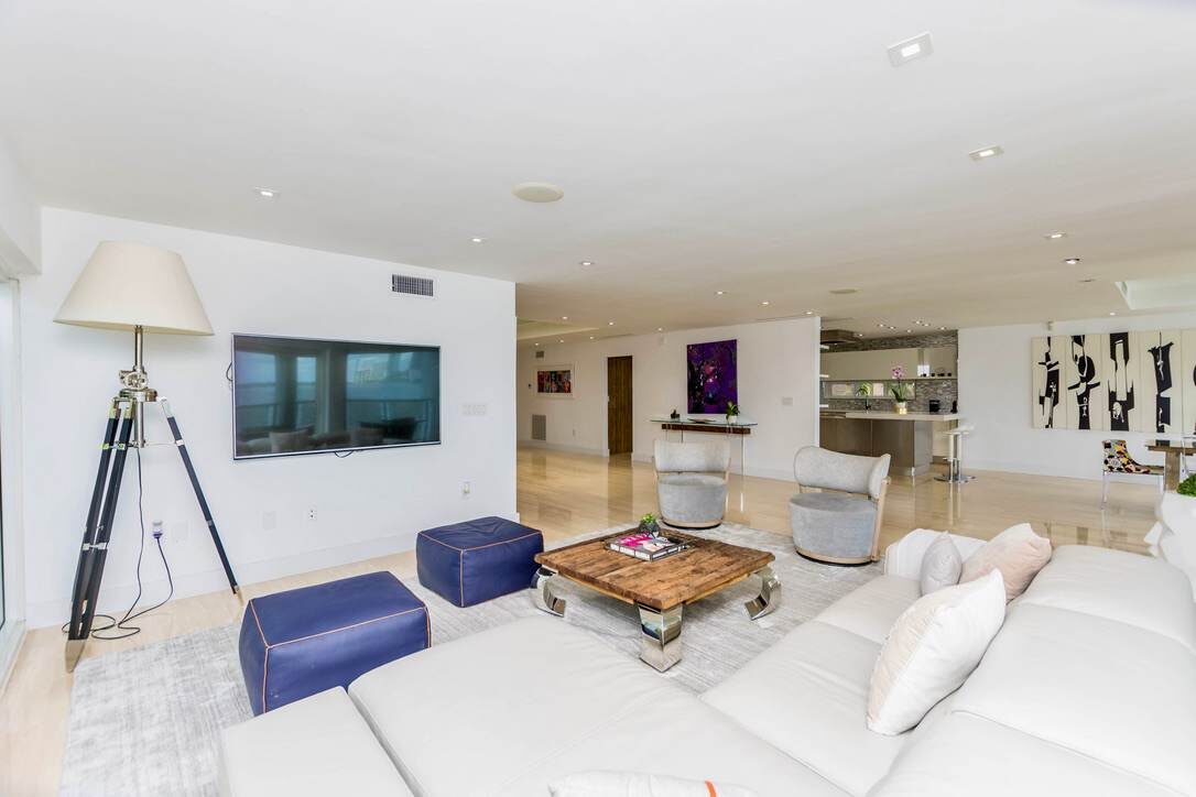 Open-concept living room featuring sectional sofa seating, lounge chairs, a wall-mounted TV, coffee table, area rug, recessed lighting, contemporary artwork, and seamless access to the adjacent kitchen and dining areas.