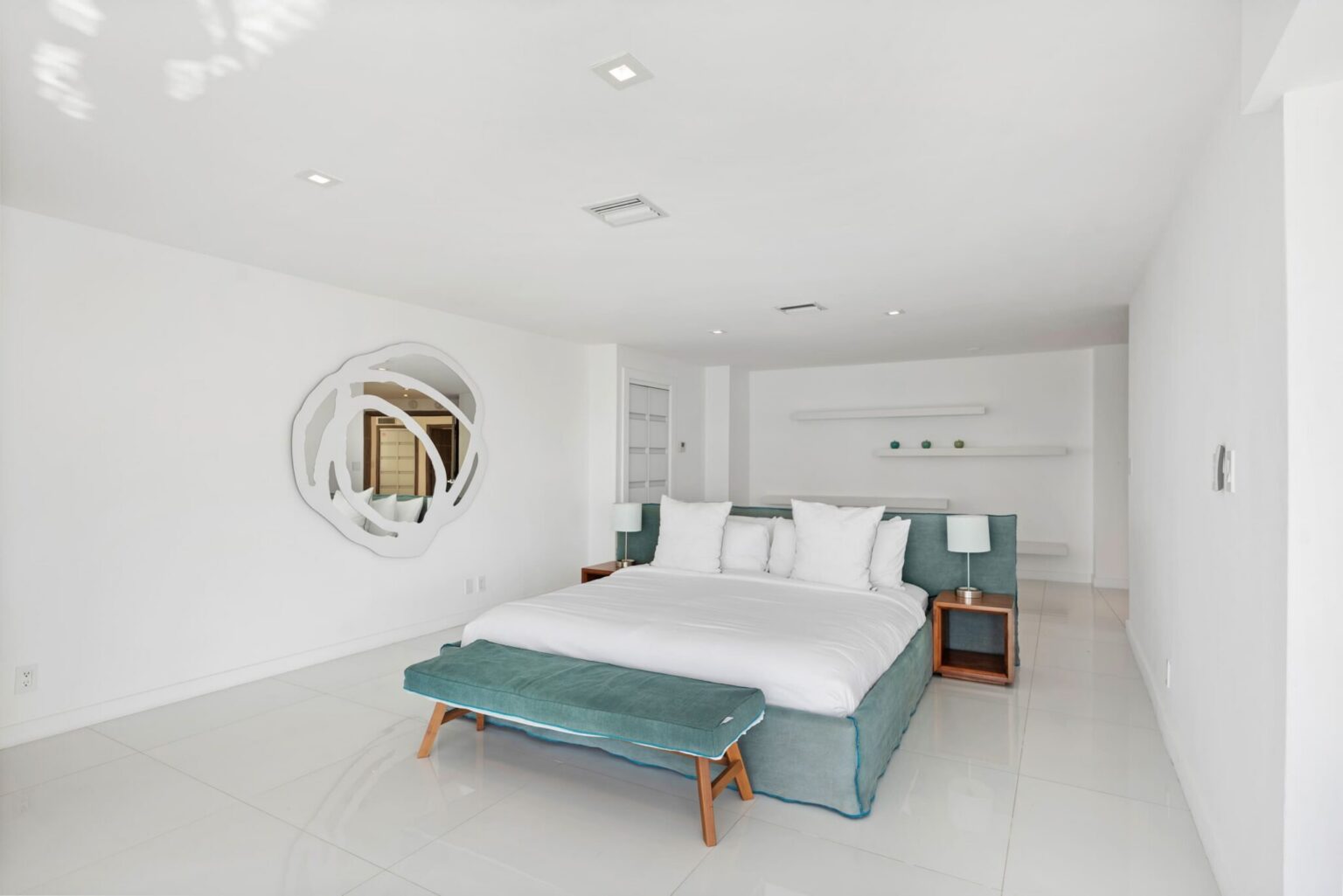 Minimalist primary suite featuring a king-size bed with premium linens, bench seating, tile flooring, bedside tables with lamps, recessed lighting, built-in shelving, and a spacious open layout designed for comfort and relaxation.