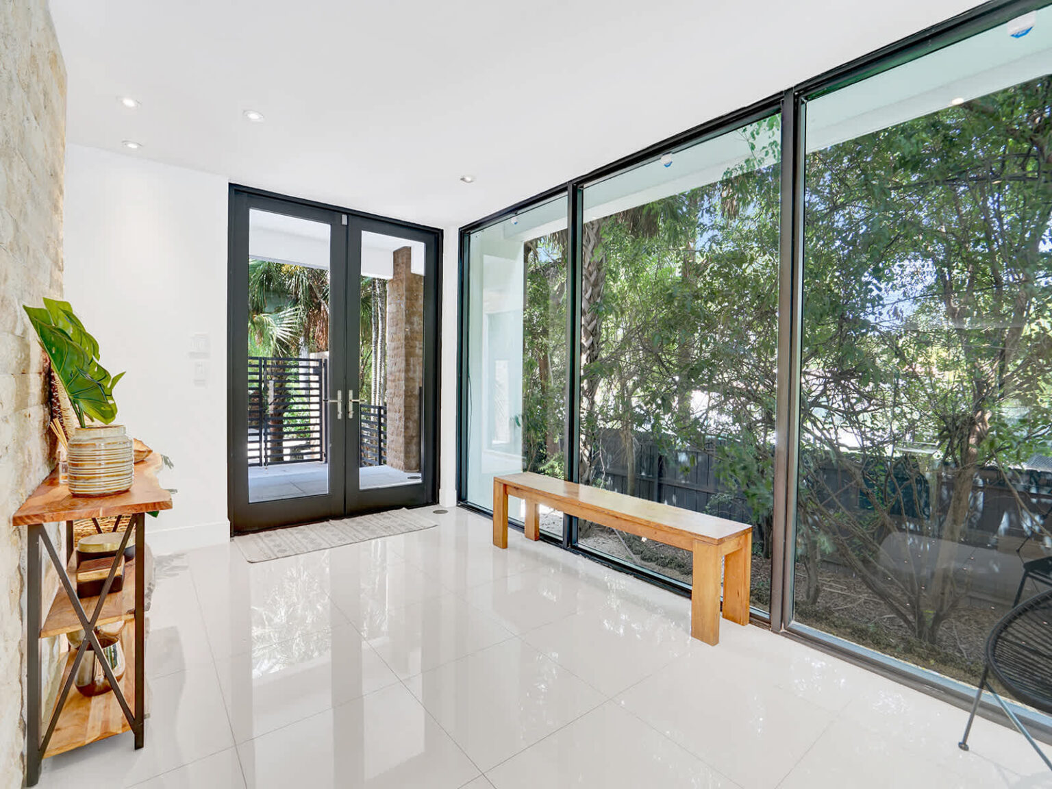 Bright corner sitting area with floor-to-ceiling windows, polished tile flooring, and tranquil views of tropical landscaping. Features a built-in bench, abundant natural light, and direct access to the outdoor patio through glass doors.